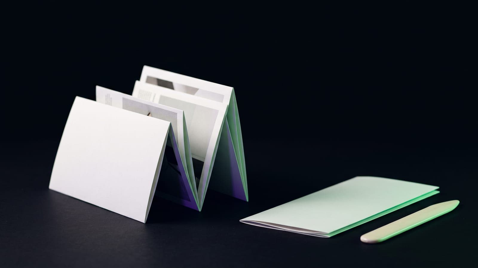 Folleto plegado en acordeón de pie sobre superficie negra, con cantos en verde menta y una plegadera de hueso al lado: la pieza detrás de cuánto cuesta un brochure o tríptico en México