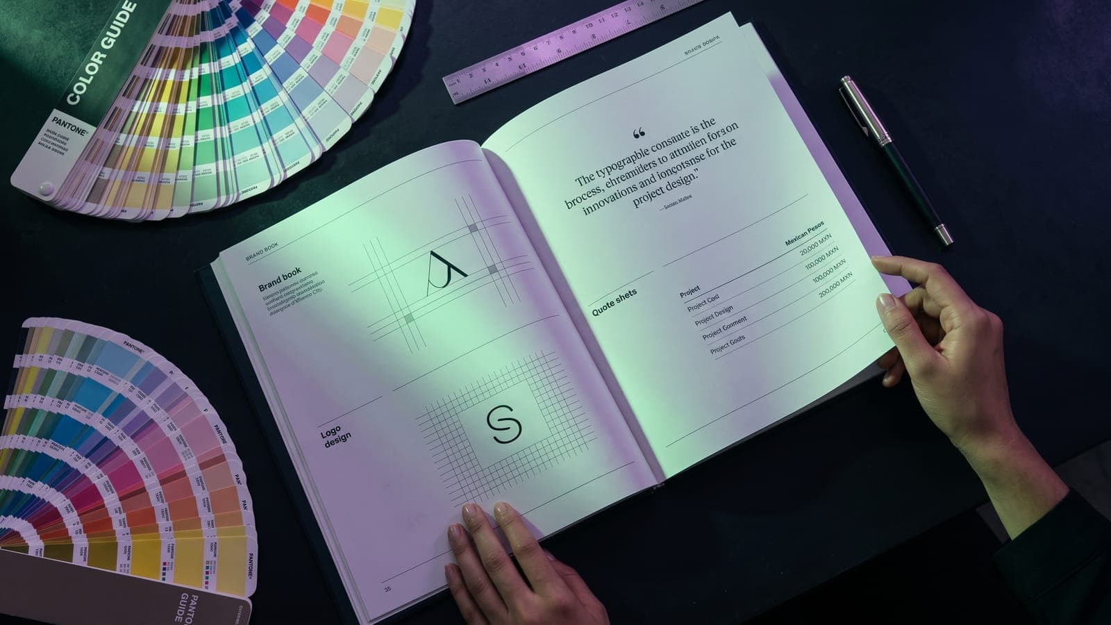 Brand book abierto con retícula de logotipo y hoja de cotización con precios en pesos mexicanos junto a abanicos Pantone, en el estudio de una agencia de branding en CDMX