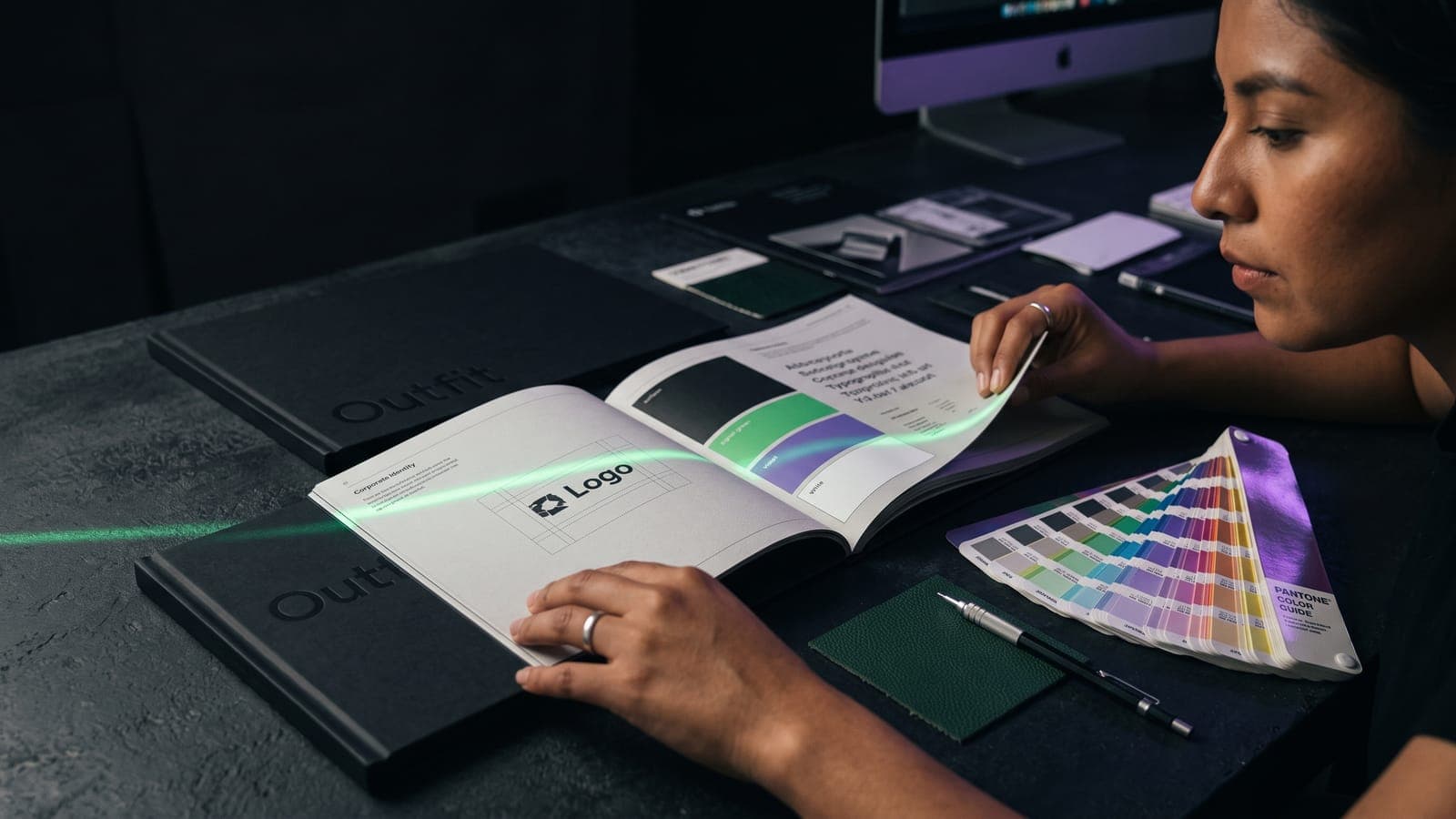 Mujer hojea un manual impreso con el logo sobre retícula y franjas de paleta en negro, verde y violeta, junto a una guía Pantone: el resultado de un buen brief de identidad corporativa