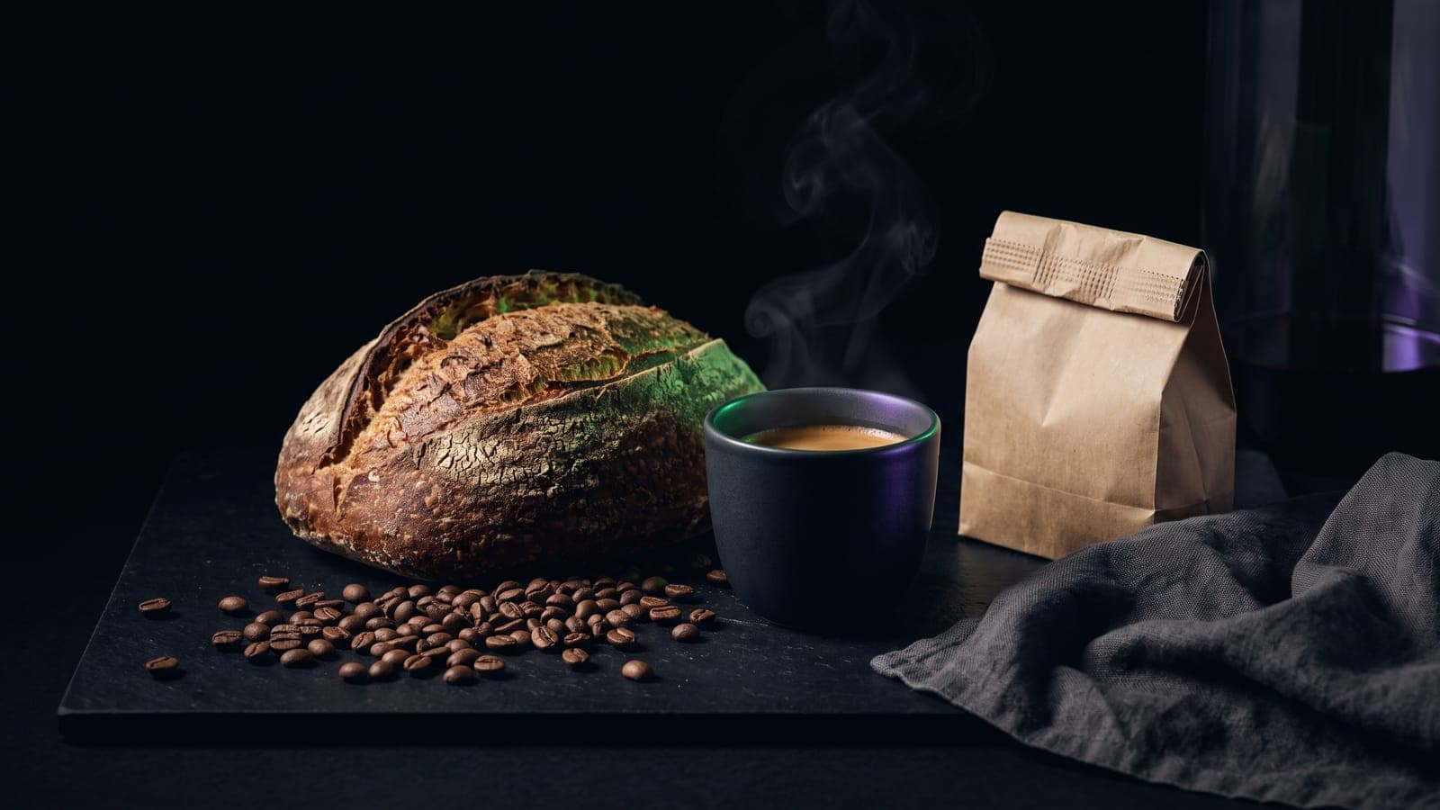 Hogaza de pan artesanal, taza negra de café humeante, granos esparcidos y una bolsa de papel kraft sobre pizarra oscura: el café, pan y repostería que ampara la clase 30 de Niza