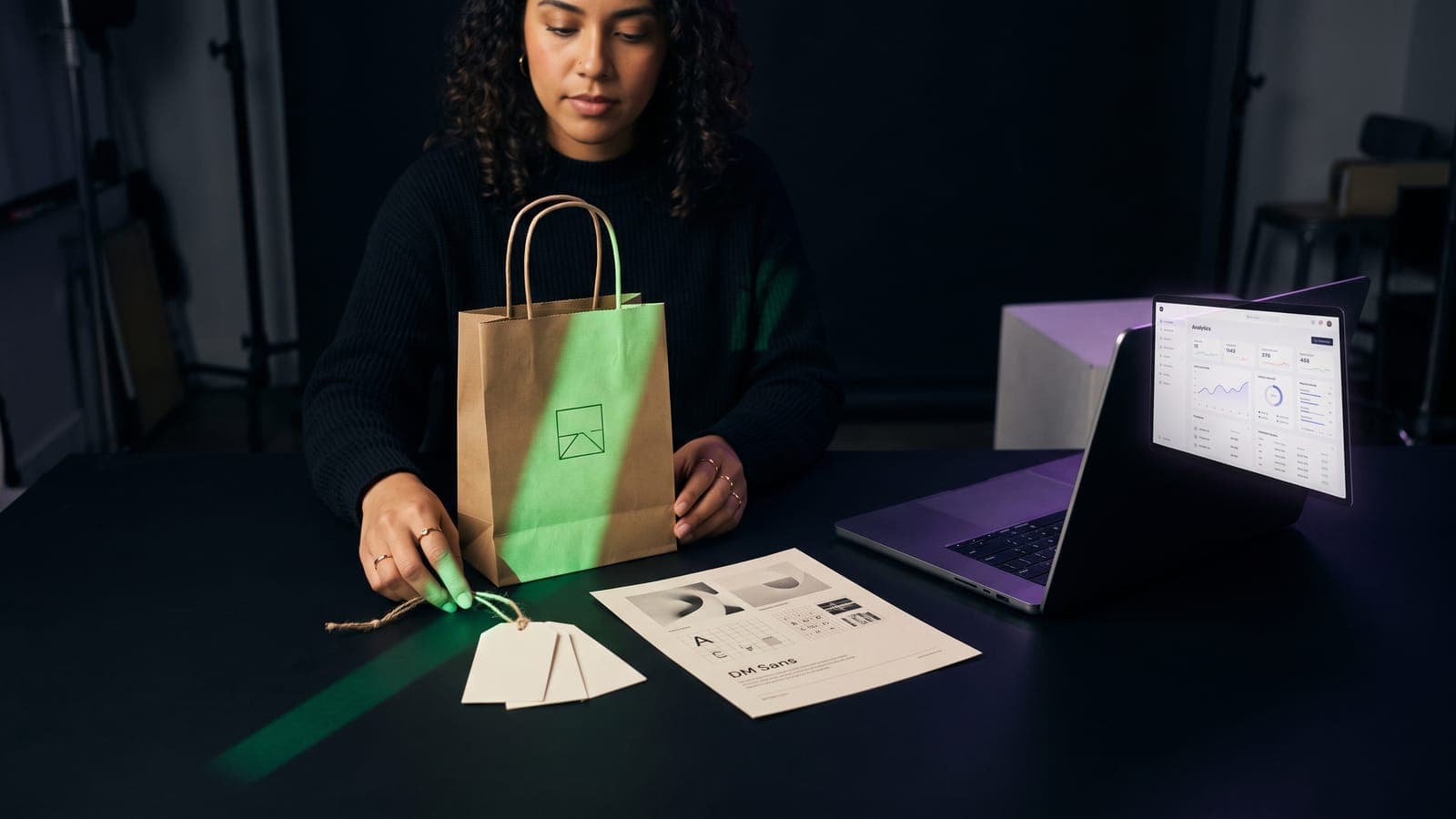 Mujer sostiene una bolsa kraft con logo minimalista junto a etiquetas colgantes y una laptop con dashboard de métricas: publicidad, gestión y retail que cubre la clase 35 de Niza