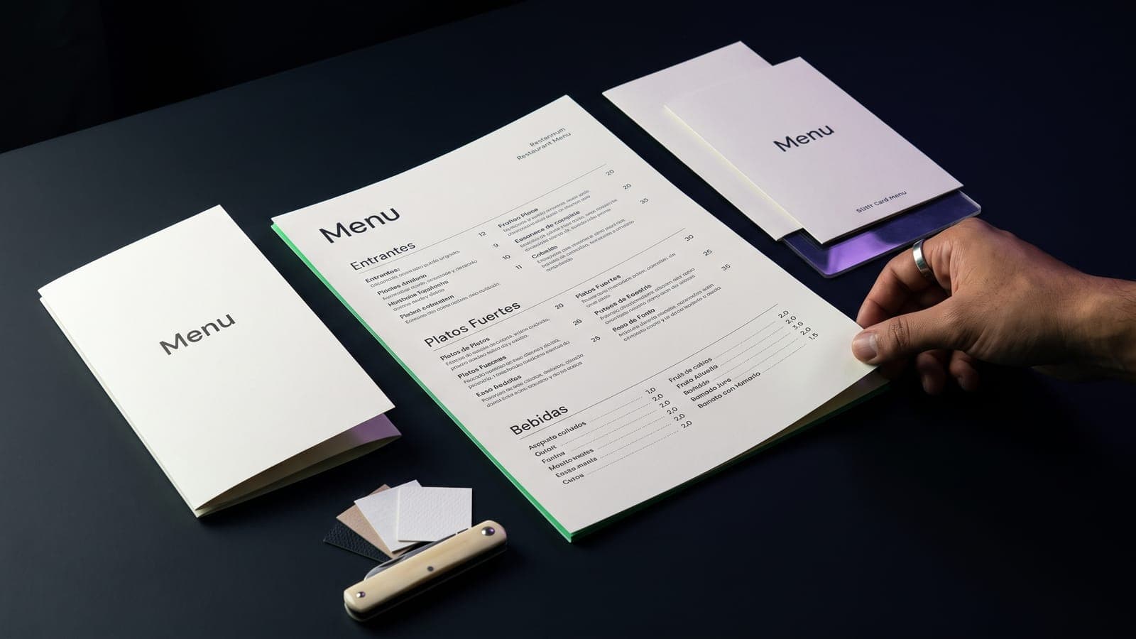 Mano sosteniendo un menú de canto verde con secciones y precios, junto a menús de tarjeta y muestras de papel sobre mesa azul noche: cuánto cuesta el diseño de un menú de restaurante