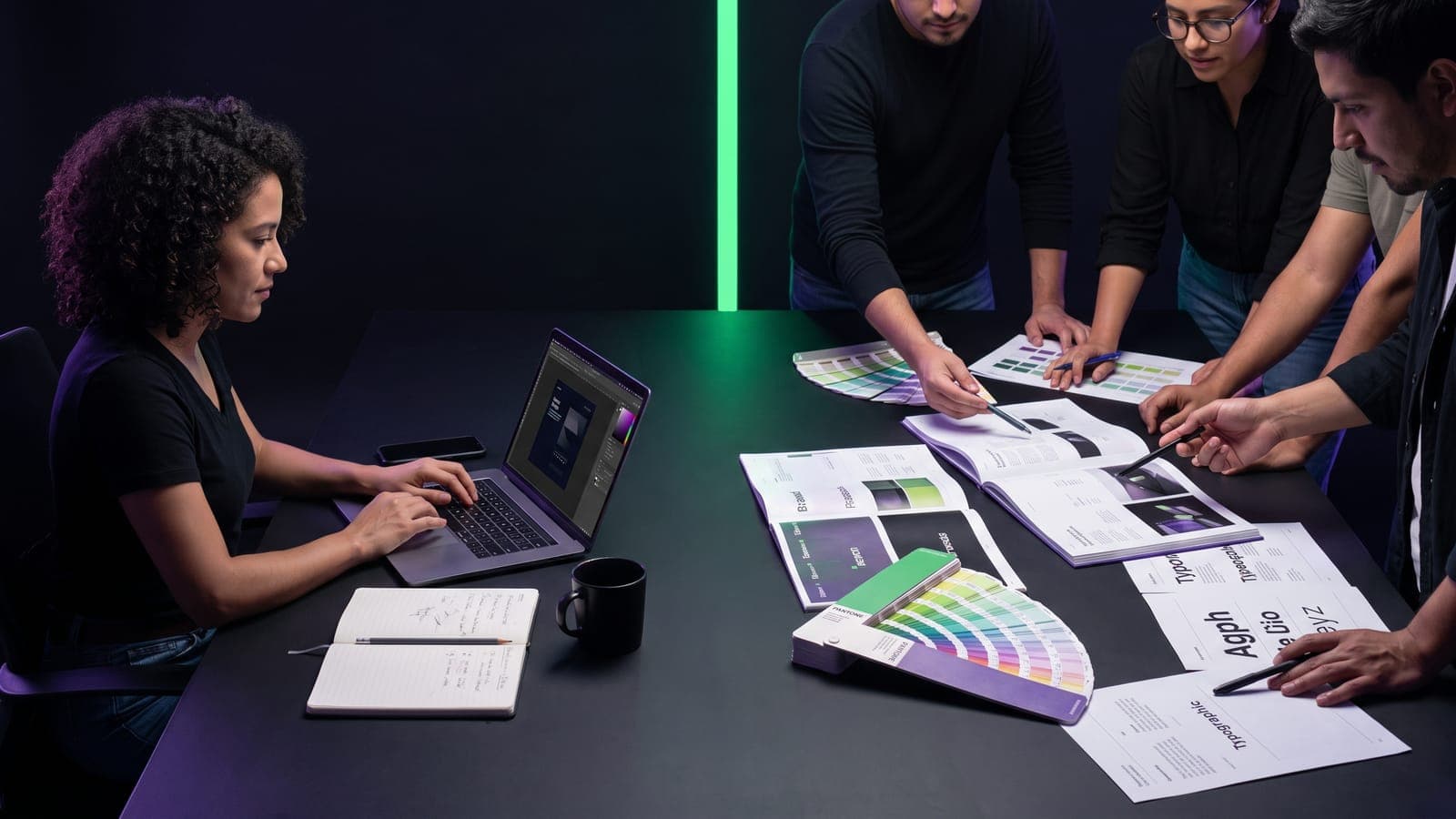 Diseñadora trabajando sola en su laptop frente a un equipo que discute sobre brand books y muestras Pantone, divididos por una línea de neón verde: freelance vs agencia de branding