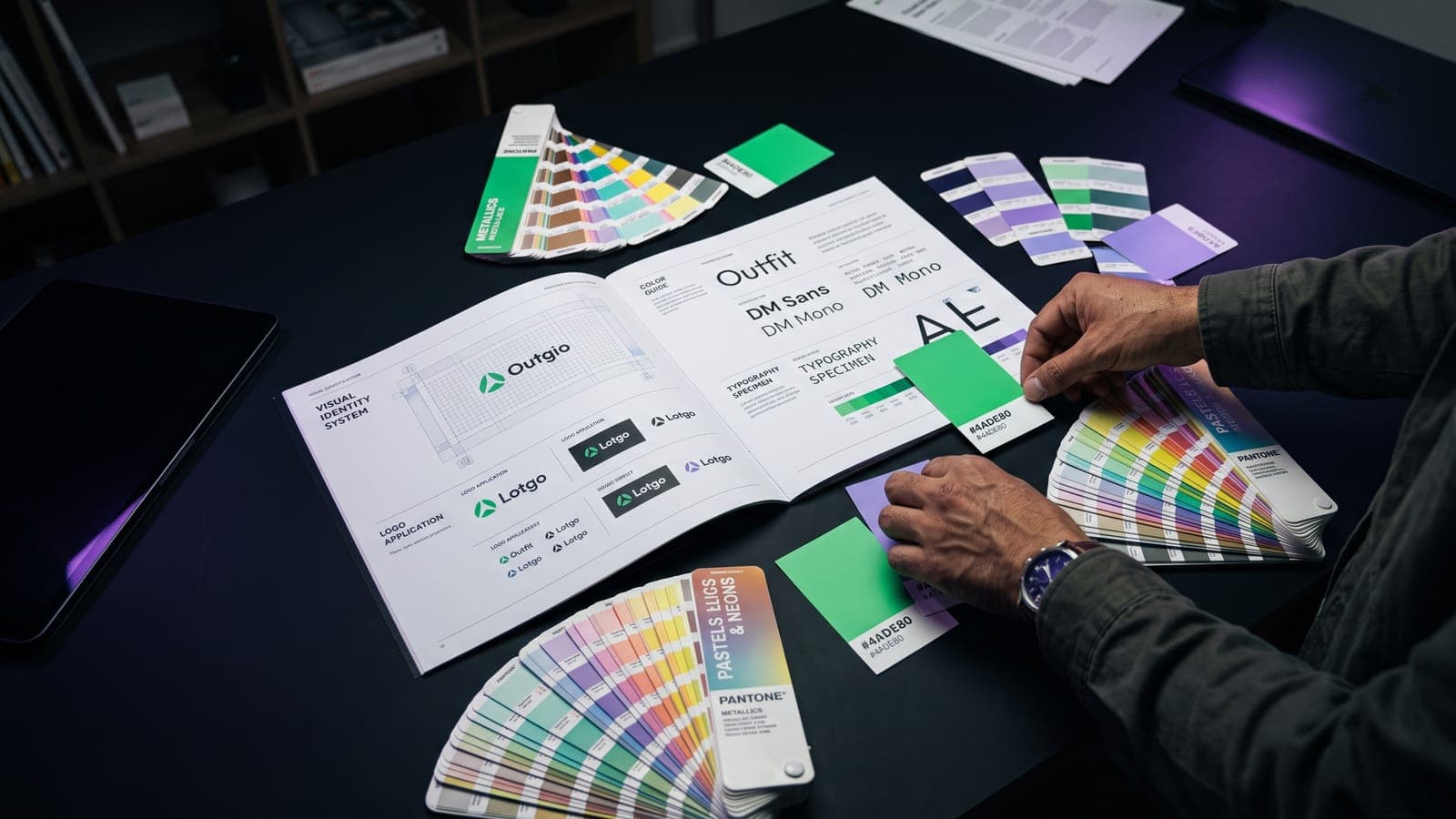Libro de sistema de identidad visual abierto con aplicaciones de logotipo, guía de color y espécimen tipográfico, rodeado de abanicos Pantone