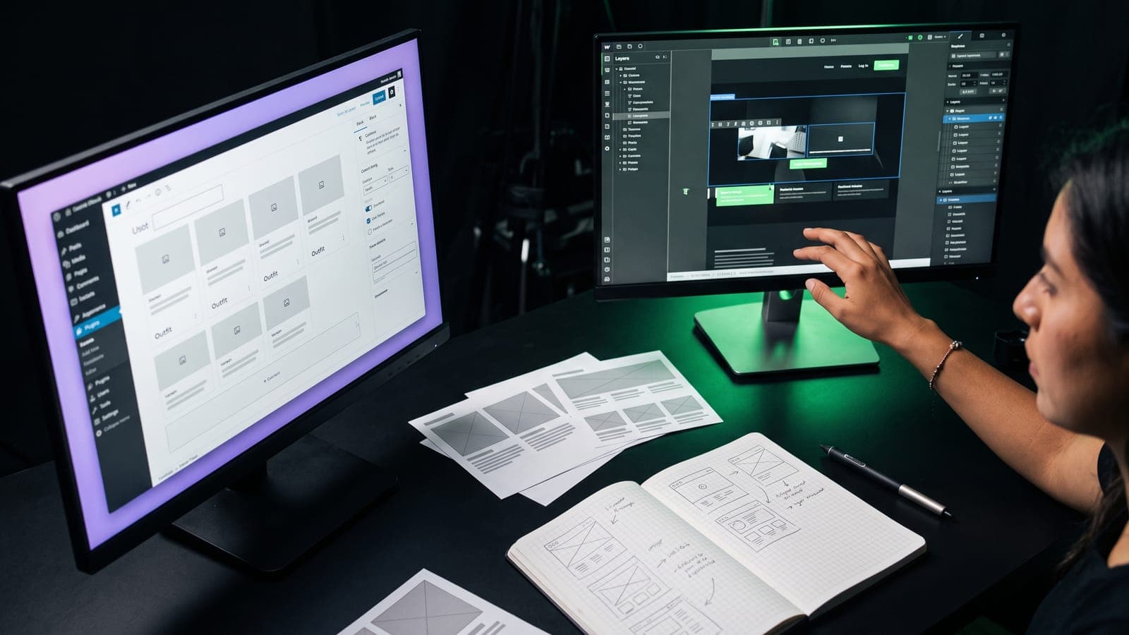 Diseñadora frente a dos monitores, uno con un editor de bloques y otro con un editor visual con panel de capas, entre wireframes impresos: la decisión WordPress vs Webflow